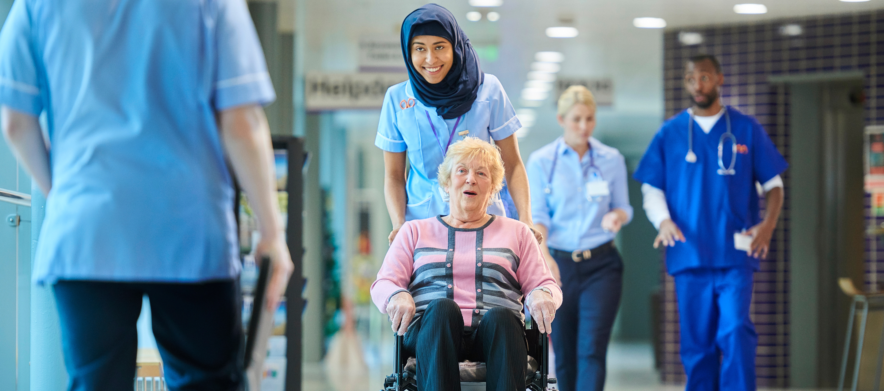 A female hospital clinician pushing an older woman in a wheelchair through a hospital corridor. Both women are smiling.