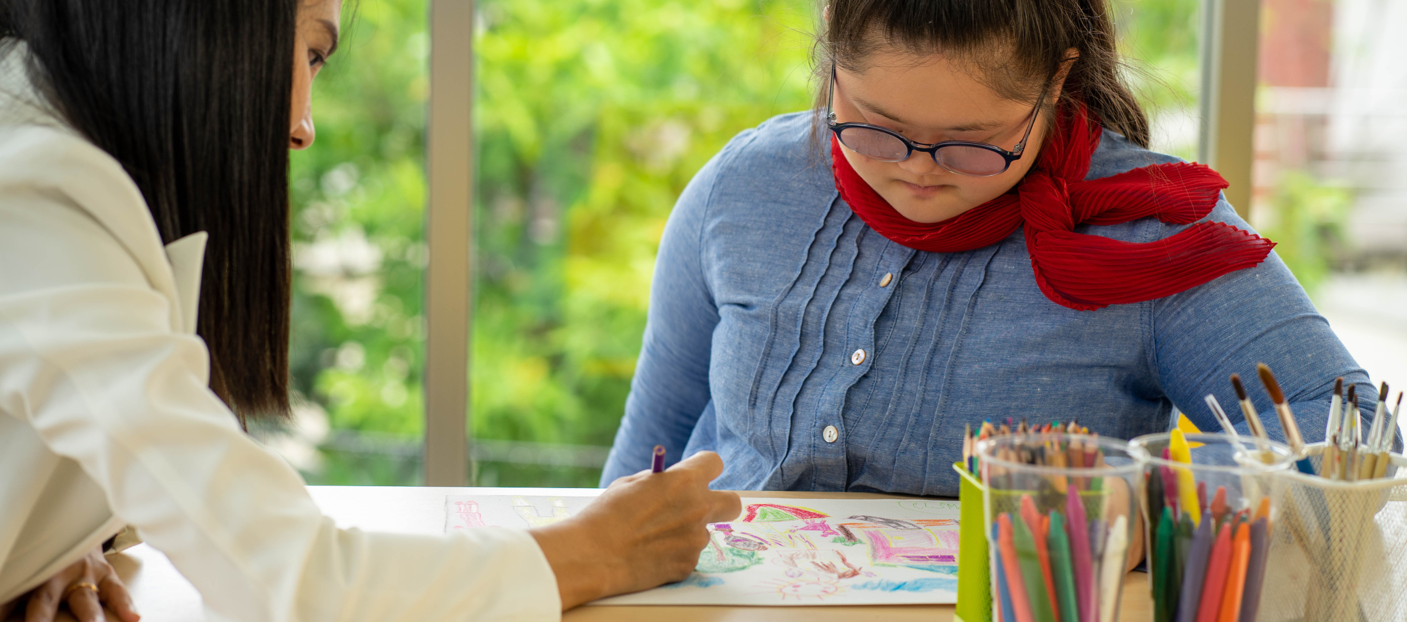 A young girl with learning disabilities drawing with a female carer sat with her.