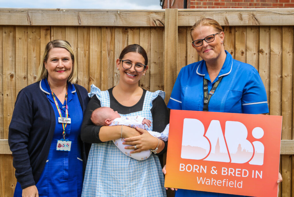 Two midwives in uniform standing in a garden with mum and baby smiling