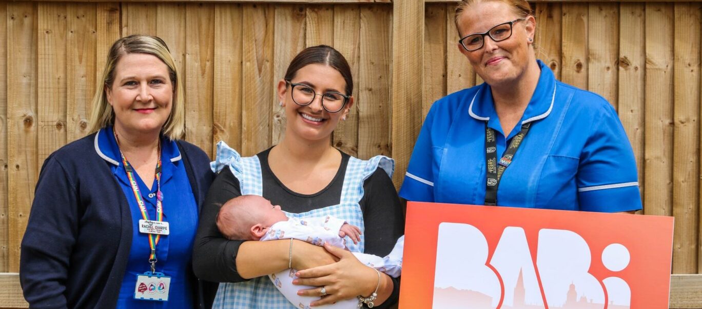 Two midwives in uniform standing in a garden with mum and baby, smiling