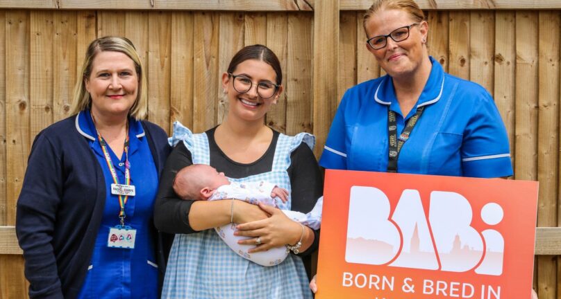 Two midwives in uniform standing in a garden with mum and baby, smiling