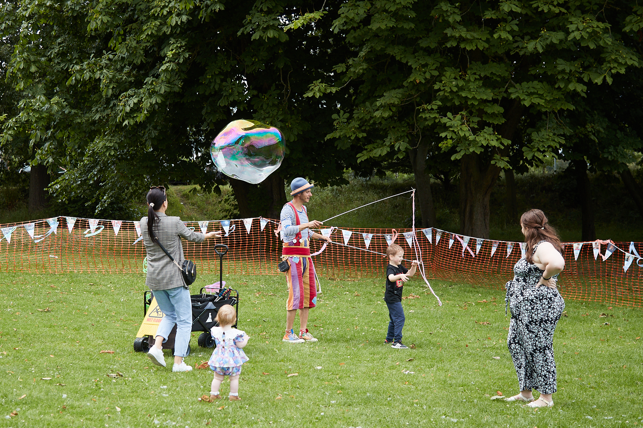 Image of a children's entertainer creating bubbles in a park. There are some children and their parents stood around him.