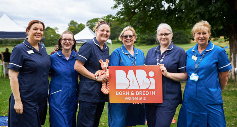 Image of a group of midwives in uniform. They are holding a BaBi Wakefield sign and a knitted orange and white bear.