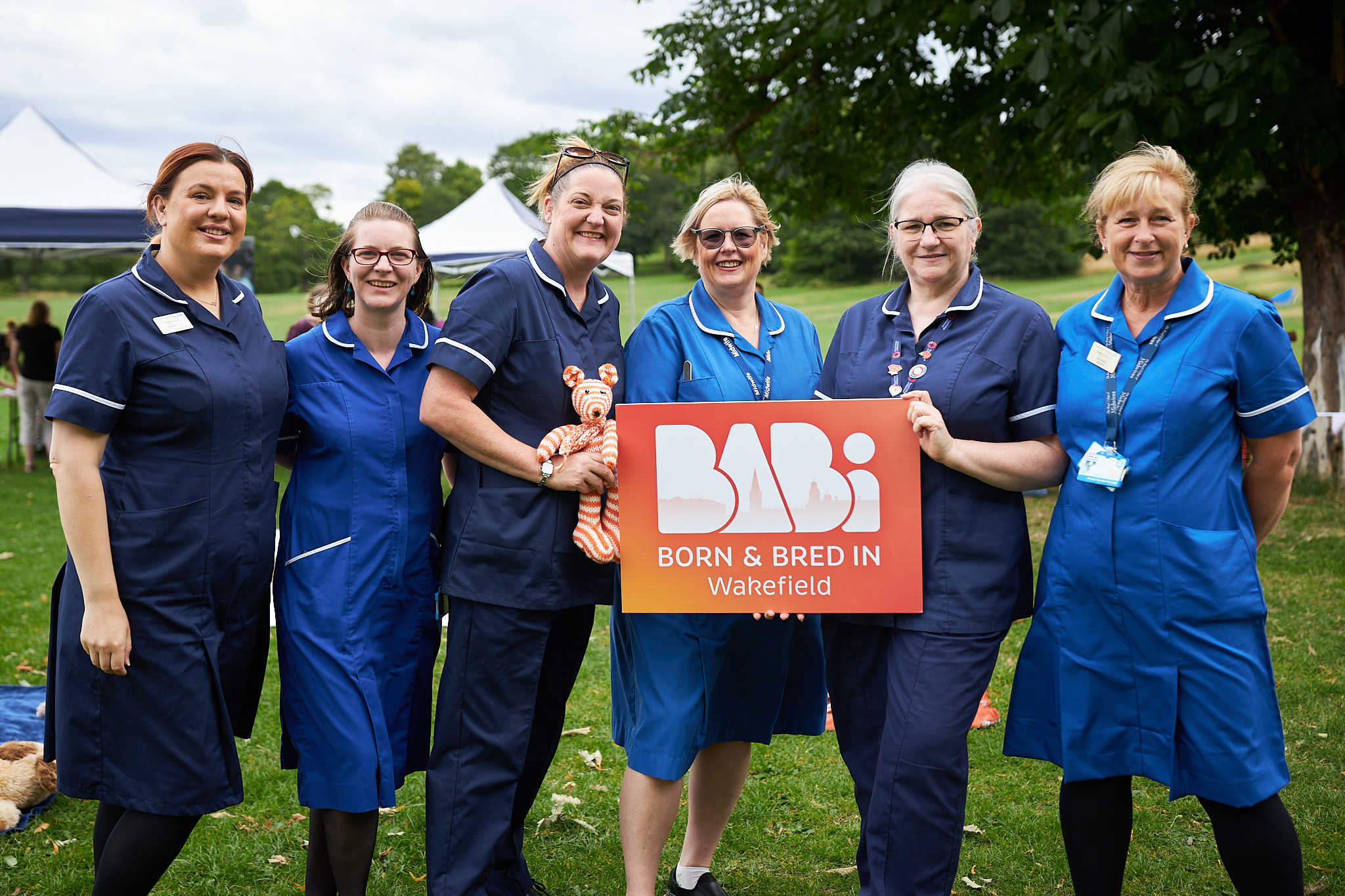 Image of a group of midwives in uniform. They are holding a BaBi Wakefield sign and a knitted orange and white bear.