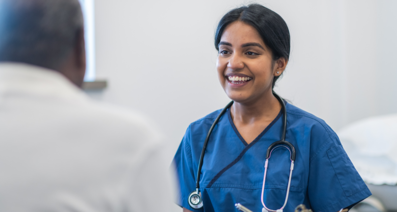 Image of a young female GP in scrubs with a stethoscope around her neck. She is also holding a clipboard and smiling at a patient.