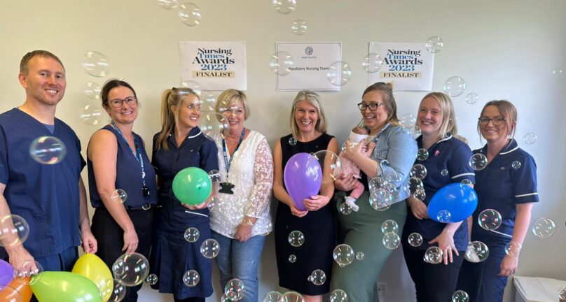Photograph of seven nurses and a GP from Health Care First Partnership smiling together will balloons and bubbles. One of the nurses is holding a baby. There is a sign on the wall behind them that says 'Nursing Times Awards 2023 finalist'. There is another sign that says ‘Health Care First Paediatric Nursing Team’.